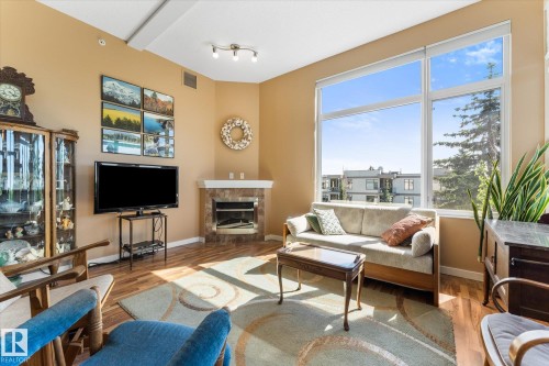 Living area featuring expansive windows, wood-finish flooring, a corner fireplace with a stone surround, and track lighting - 408 11140 68 Avenue, Edmonton, AB 