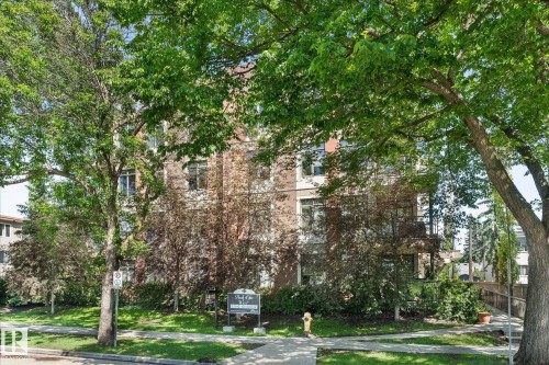 Brick and stucco multi-story building featuring mature trees, landscaped grounds, a paved sidewalk, and a dedicated entrance sign - 408 11140 68 Avenue, Edmonton, AB 