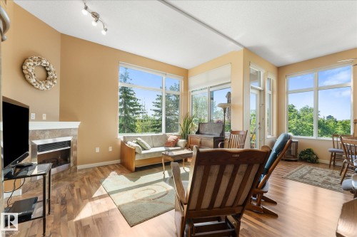 Spacious living area featuring expansive windows, wood-finish flooring, a corner fireplace with a tiled surround and mantel, and track lighting - 408 11140 68 Avenue, Edmonton, AB 