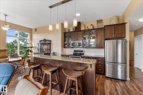 Open-concept kitchen featuring wood-finish flooring, stainless steel appliances, a breakfast bar with stone countertops, pendant lighting, and ample cabinetry - 408 11140 68 Avenue, Edmonton, AB 