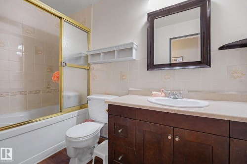 Bathroom featuring a full vanity with a white basin sink, framed mirror, and built-in bathtub with a gold-toned sliding shower door - 10242 161 Street, Edmonton, AB 