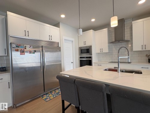 Contemporary kitchen featuring an oversized island with an undermount sink, a subway tile backsplash, stainless steel appliances, white cabinetry, and wood-finish flooring - 9952 227 Street, Edmonton, AB 