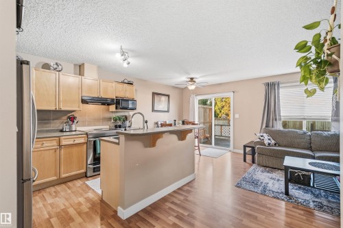 Open-concept living space featuring wood-finish flooring, a kitchen island with seating, stainless steel appliances, and wood cabinetry - 21207 90Ave, Edmonton, AB 