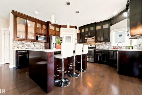 Kitchen featuring wood-finish flooring, two-tone cabinetry with glass-front uppers, a mosaic tile backsplash, a kitchen island with pendant lighting, and stainless steel appliances - 5110 Rue Eaglemont, Beaumont, AB 