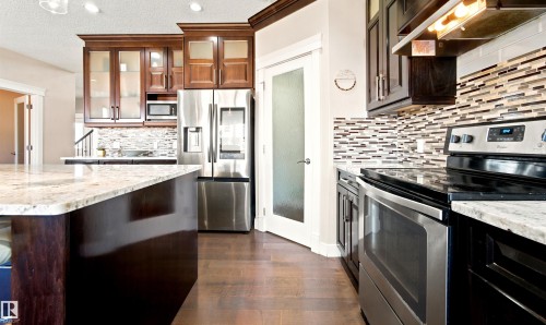 Gourmet kitchen featuring a large island with a light-toned countertop, stainless steel appliances, dark wood-finish cabinetry, and a mosaic tile backsplash - 5110 Rue Eaglemont, Beaumont, AB 