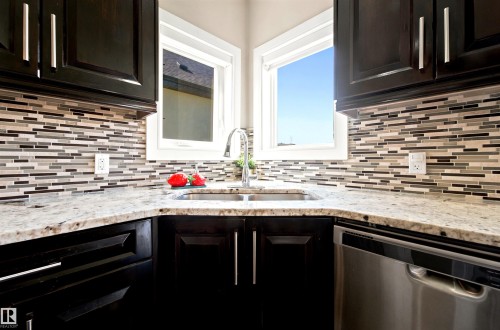 Kitchen corner featuring dark wood-finish cabinetry with brushed nickel hardware, light-toned stone countertops, and a full-height mosaic tile backsplash - 5110 Rue Eaglemont, Beaumont, AB 