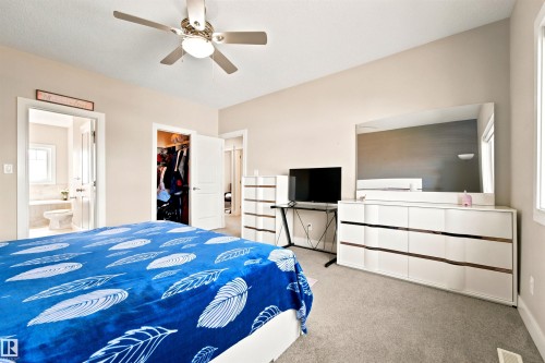 Carpeted room featuring a ceiling fan with integrated lighting, a walk-in closet, a bathroom entry, and a window providing natural light - 5110 Rue Eaglemont, Beaumont, AB 