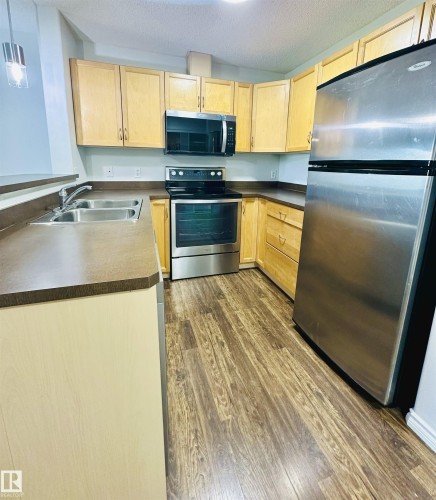 Kitchen featuring wood-finish flooring, light wood cabinetry, dark countertops, and stainless steel appliances including a refrigerator, range, and microwave - 318 646 Mcallister Loop, Edmonton, AB 