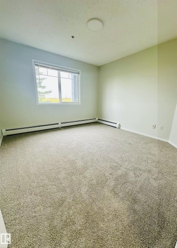 Spacious room featuring neutral carpeting, a large window, white baseboard heating, and a flush-mount ceiling light fixture - 318 646 Mcallister Loop, Edmonton, AB 
