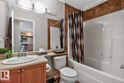 Bathroom featuring a built-in bathtub with shower, a vanity with a drop-in sink, wood-finish cabinetry, and a large mirror with integrated lighting - 320 5005 165 Avenue, Edmonton, AB - Indoor Photo Showing Bathroom