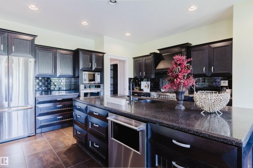 Modern kitchen featuring dark wood cabinetry, stainless steel appliances, a large center island with a dark speckled countertop, and a black subway tile backsplash - 3438 West Landing Landing, Edmonton, AB 