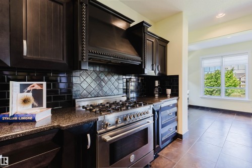 Gourmet kitchen featuring dark wood cabinetry, a stainless steel range with a custom hood, black speckled countertops, and a black subway tile backsplash with a decorative inset - 3438 West Landing Landing, Edmonton, AB 