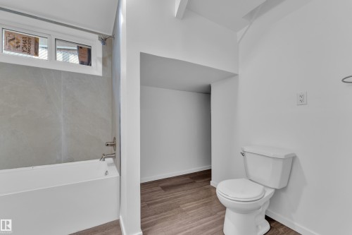 Bathroom featuring a white bathtub with light gray tile surround, chrome fixtures, a white toilet, and wood-finish flooring - 8104 135 Avenue, Edmonton, AB 