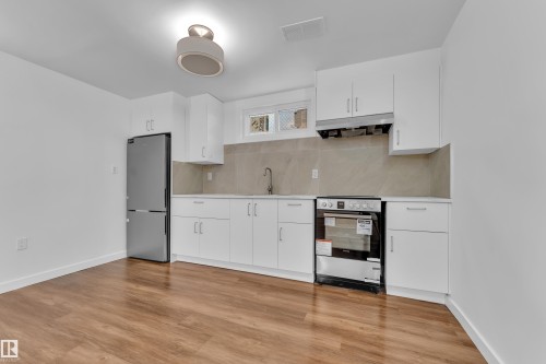 Modern kitchen featuring white cabinetry, stainless steel appliances, a light-toned backsplash, wood-finish flooring, and a contemporary ceiling fixture - 8104 135 Avenue, Edmonton, AB 