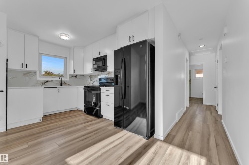 Modern kitchen featuring white cabinetry, black hardware, black appliances, a white and gray patterned backsplash, and wood-finish flooring - 8104 135 Avenue, Edmonton, AB 