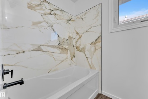 Bathtub featuring large-format porcelain tile surround with marble-look veining, matte black fixtures, and a white framed window - 8104 135 Avenue, Edmonton, AB 