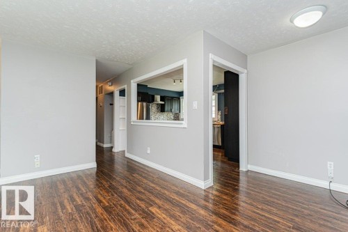 Spacious room with wood-finish flooring, light grey walls, white baseboards, and a pass-through window to the kitchen - 13411 140 Street, Edmonton, AB 