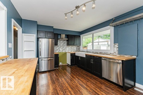 Modern kitchen featuring black cabinetry, stainless steel appliances, wood-finish countertops, a farmhouse sink, and a mosaic tile backsplash - 13411 140 Street, Edmonton, AB 