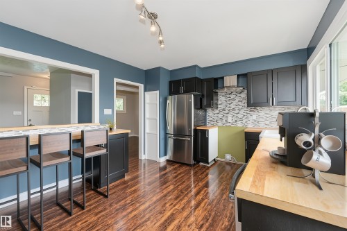 Contemporary kitchen featuring blue accent walls, dark wood-finish flooring, black cabinetry, butcher block countertops, and a mosaic tile backsplash - 13411 140 Street, Edmonton, AB 