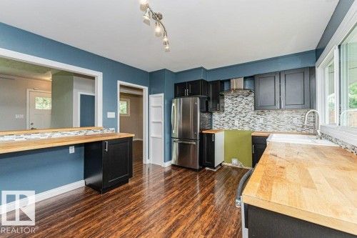 Kitchen featuring wood-finish flooring, butcher block countertops, stainless steel refrigerator, black cabinetry, and mosaic tile backsplash - 13411 140 Street, Edmonton, AB 