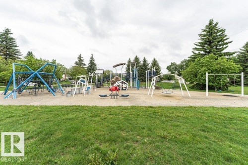 Outdoor playground featuring multiple play structures, swings, and a red seesaw, all set on a sand base with surrounding green grass and mature trees - 13411 140 Street, Edmonton, AB 