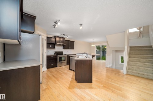 Open-concept kitchen featuring dark wood cabinetry, stainless steel appliances, a center island with a built-in sink, and light wood-finish flooring - 11312 13 Ave Sw, Edmonton, AB 