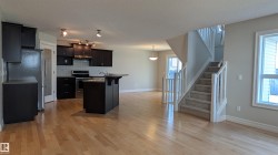 Open-concept living area featuring wood-finish flooring, a kitchen with dark cabinetry and stainless steel appliances, a central kitchen island with a sink, and a carpeted staircase with white railings - 