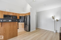 Open-concept kitchen featuring light wood cabinetry, dark countertops, dark mosaic tile backsplash, and stainless steel appliances, all complemented by wood-finish flooring and a vaulted ceiling - 