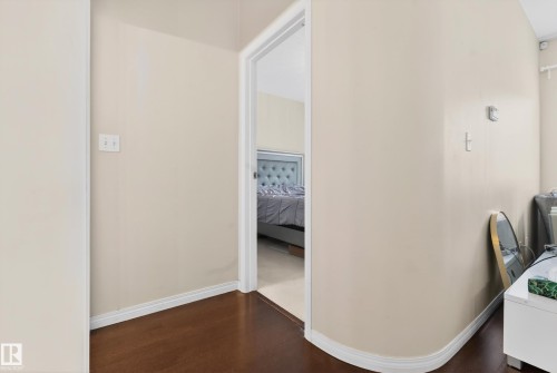 Hallway with wood-finish flooring and light-toned walls leading to a room with carpeted flooring and a tufted headboard - 226 4304 139 Avenue, Edmonton, AB 