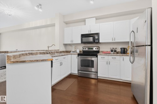 Kitchen featuring white cabinetry, light granite-finish countertops, stainless steel appliances, a corner sink with a high-arch faucet, and wood-finish flooring - 226 4304 139 Avenue, Edmonton, AB 