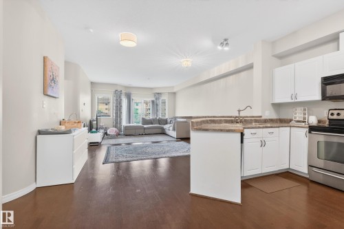 Open-concept living space featuring wood-finish flooring, a kitchen with white cabinetry and stainless steel appliances, and a built-in kitchen island with an undermount sink - 226 4304 139 Avenue, Edmonton, AB 