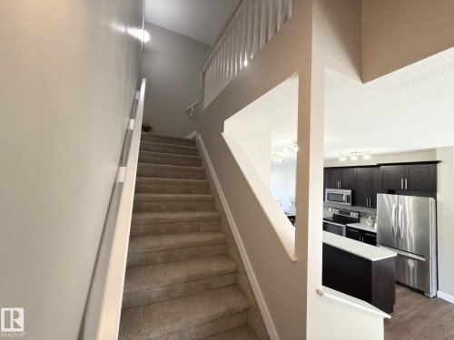 Carpeted staircase with white railings, offering a partial view of a kitchen featuring dark cabinetry, stainless steel appliances, and wood-finish flooring - 3110 Paisley Road, Edmonton, AB 