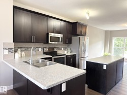 Kitchen featuring dark wood-finish cabinetry, light-toned countertops, a stainless steel double-basin sink, and a tile backsplash with mosaic accents - 