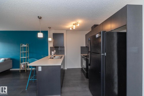 Contemporary kitchen featuring dark cabinetry, a breakfast bar with pendant lighting, integrated sink, black appliances, and dark wood-finish flooring - 431 344 Windermere Road, Edmonton, AB - Indoor Photo Showing Kitchen With Double Sink