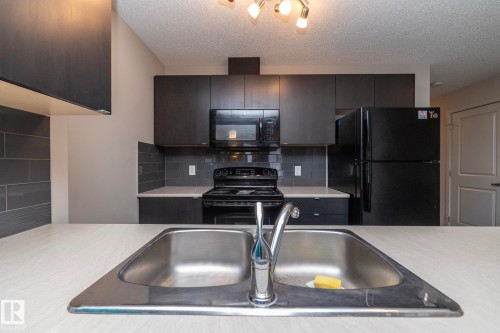 Modern kitchen featuring dark cabinetry, stainless steel double basin sink with chrome faucet, black appliance suite, light-toned countertops, and a gray subway tile backsplash - 431 344 Windermere Road, Edmonton, AB - Indoor Photo Showing Kitchen With Double Sink