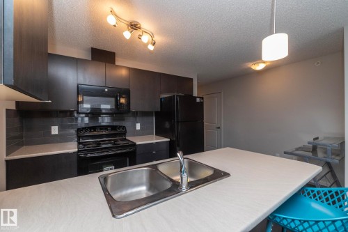 Kitchen featuring a double basin stainless steel sink, dark wood-finish cabinetry, a black appliance package, and a subway tile backsplash - 431 344 Windermere Road, Edmonton, AB - Indoor Photo Showing Kitchen With Double Sink