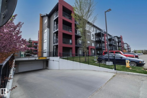Modern multi-story building featuring a red and gray facade, multiple balconies with black railings, a sloped entrance to an underground parking garage, and a surface parking lot - 431 344 Windermere Road, Edmonton, AB - Outdoor With Balcony With Facade