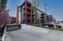 Modern multi-story building featuring a red and gray facade, multiple balconies with black railings, a sloped entrance to an underground parking garage, and a surface parking lot - 431 344 Windermere Road, Edmonton, AB  - Outdoor With Balcony With Facade 