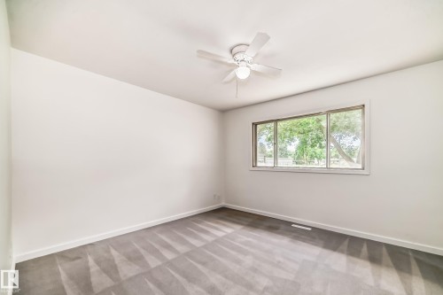Bright interior room featuring a large window with white trim, a ceiling fan with light fixture, light gray carpet, and white baseboards - 11937 39 Street Nw, Edmonton, AB 