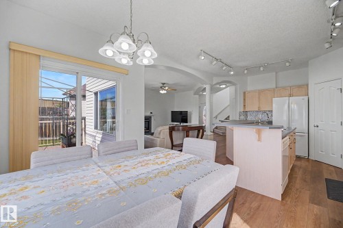 Dining area featuring a contemporary chandelier, sliding glass door with vertical blinds, wood-finish flooring, and open access to the kitchen with light wood cabinetry and a neutral countertop - 262 Dunvegan Road, Edmonton, AB 