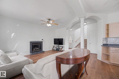 Spacious interior featuring a fireplace with a dark stone surround, wood-finish flooring, a ceiling fan with integrated lighting, and a partial kitchen area with light cabinetry and a patterned tile backsplash - 262 Dunvegan Road, Edmonton, AB 