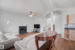 Spacious interior featuring a fireplace with a dark stone surround, wood-finish flooring, a ceiling fan with integrated lighting, and a partial kitchen area with light cabinetry and a patterned tile backsplash - 