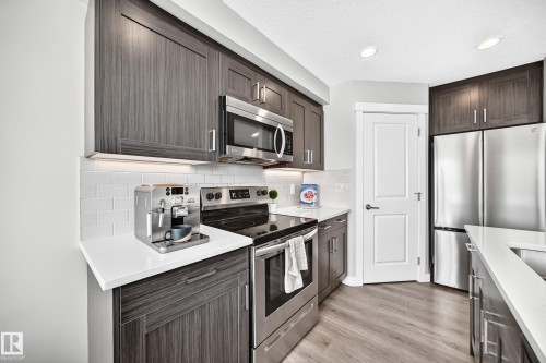 Modern kitchen featuring wood-grain cabinetry, white countertops, a subway tile backsplash, stainless steel appliances, and wood-finish flooring - 93 Richmond Link, Fort Saskatchewan, AB 