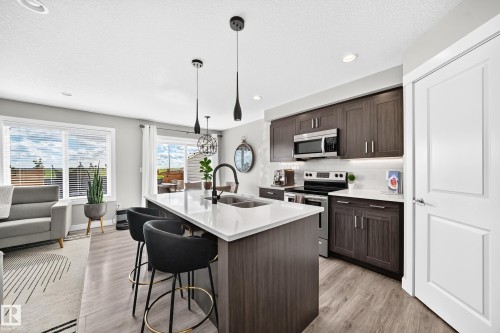 Contemporary kitchen featuring a large island with seating, pendant lighting, stainless steel appliances, dark wood-finish cabinetry, and light-colored countertops - 93 Richmond Link, Fort Saskatchewan, AB 