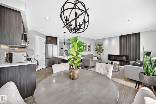 Open-concept living space featuring a contemporary linear fireplace, dark wood-finish cabinetry, stainless steel appliances, and a modern spherical chandelier - 93 Richmond Link, Fort Saskatchewan, AB 