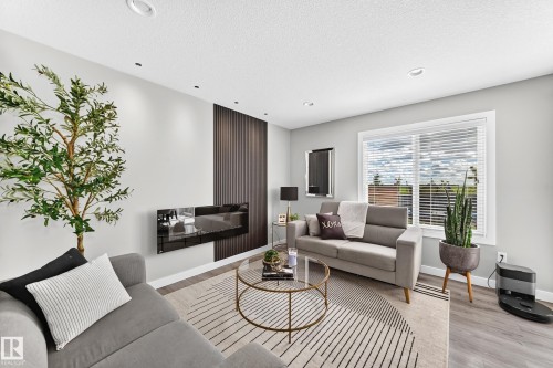 Living area featuring a modern wall-mounted fireplace, a dark wood-paneled accent wall, recessed lighting, wood-finish flooring, and a large window with horizontal blinds - 93 Richmond Link, Fort Saskatchewan, AB 