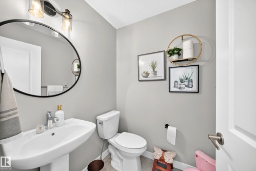 Powder room featuring a pedestal sink with chrome faucet, round black-framed mirror, and a two-light vanity fixture with clear glass shades - 93 Richmond Link, Fort Saskatchewan, AB 