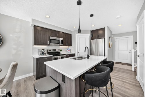 Modern kitchen featuring dark wood-finish cabinetry, stainless steel appliances, a white tile backsplash, and a large island with a dual sink and pendant lighting - 93 Richmond Link, Fort Saskatchewan, AB 