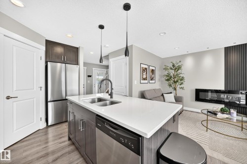 Open-concept kitchen featuring a large island with white countertop, integrated stainless steel sink, and dishwasher - 93 Richmond Link, Fort Saskatchewan, AB 