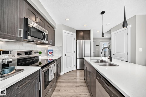 Contemporary kitchen featuring a large island with dual basin sink and matte black faucet, stainless steel appliances, wood-finish cabinetry, white subway tile backsplash, and recessed lighting - 93 Richmond Link, Fort Saskatchewan, AB 